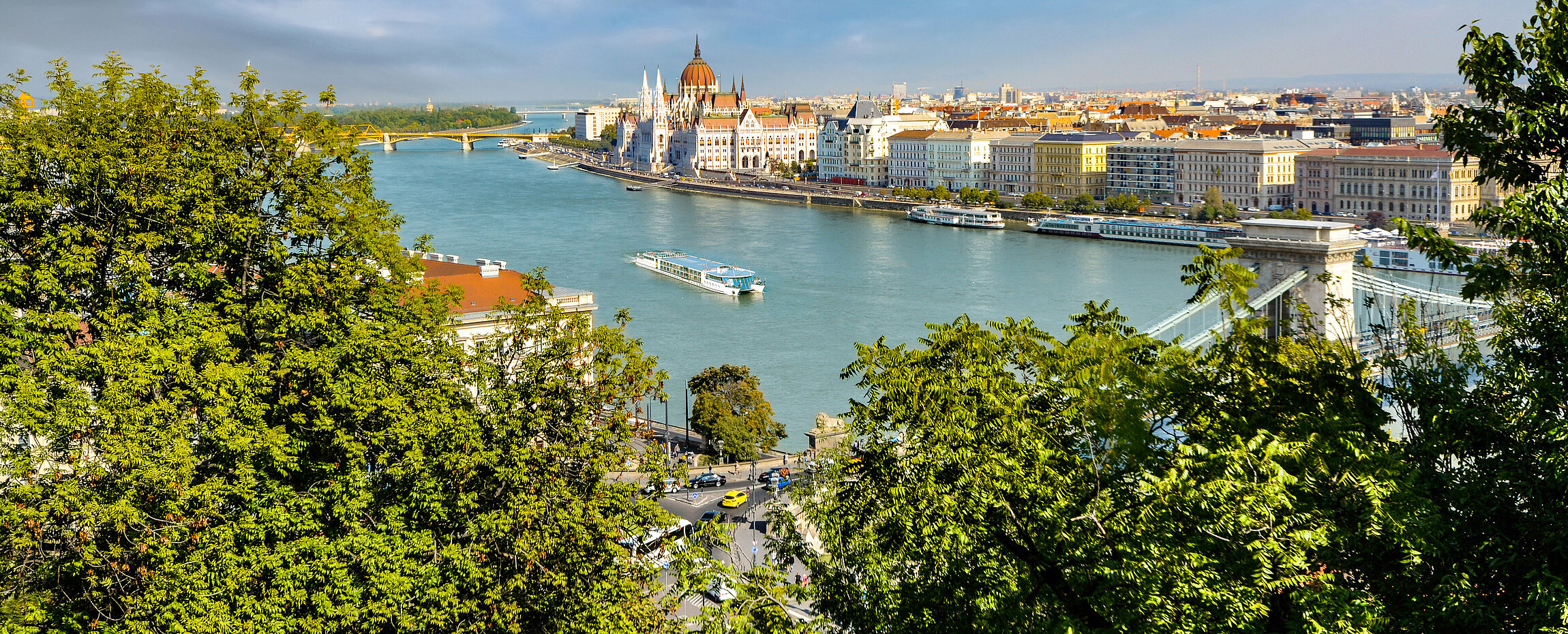 Luftaufnahme der Donau in Budapest mit Blick auf das ungarische Parlamentsgebäude und die Kettenbrücke, umrahmt von grünen Baumwipfeln.