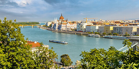 Luftaufnahme der Donau in Budapest mit Blick auf das ungarische Parlamentsgebäude und die Kettenbrücke, umrahmt von grünen Baumwipfeln.